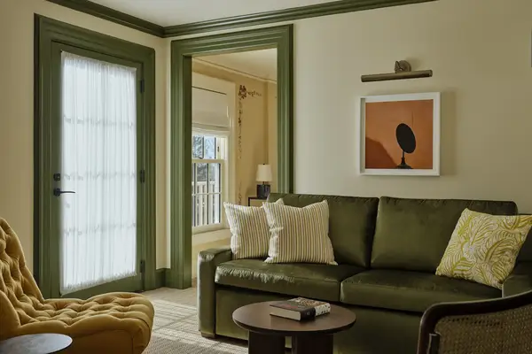 A cozy one-bedroom suite living room with a sofa, throw pillows, an armchair, a framed artwork on the wall, and a door leading to another area in the background