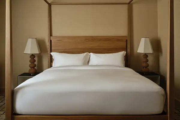 Wooden bed with white bedding, flanked by two side tables with lamps, set against a beige wall