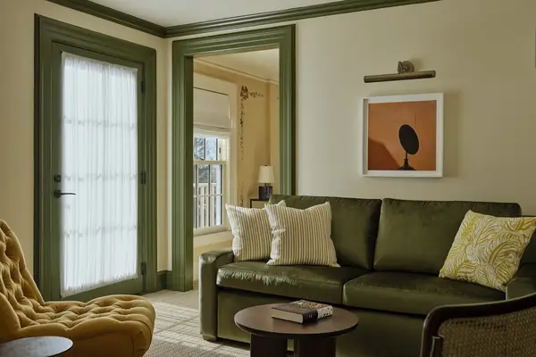 A cozy one-bedroom suite living room with a sofa, throw pillows, an armchair, a framed artwork on the wall, and a door leading to another area in the background