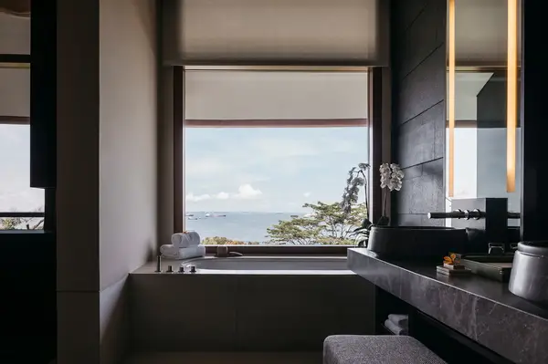 Luxury bathroom with a view of the sea through a large window, featuring a counter with a sink and a towel rack next to the bathtub