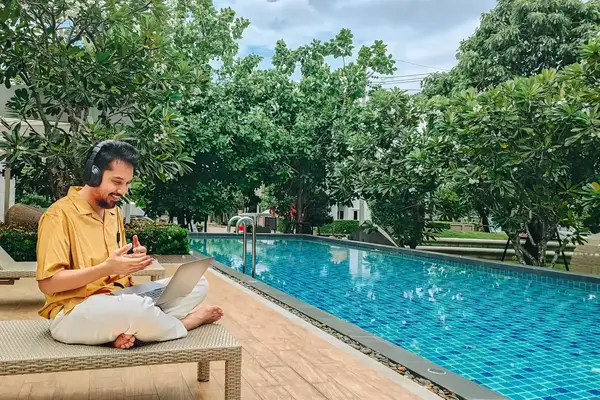Freelancer working with laptop and sitting on deck chair near the pool at resort on summer vacation.