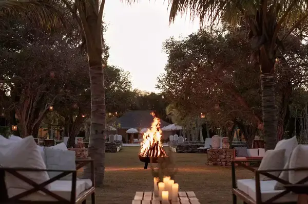 Outdoor scene featuring a lit firepit, surrounded by seating and palm trees, with additional decor in the background