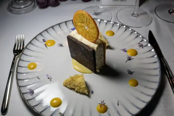 Gourmet dessert plated with cream, cake pieces, and a dried orange slice for garnish, surrounded by sauce drops and floral accents