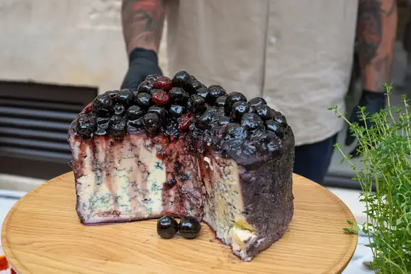 A wheel of cheese topped with a generous amount of dark berries displayed on a wooden board