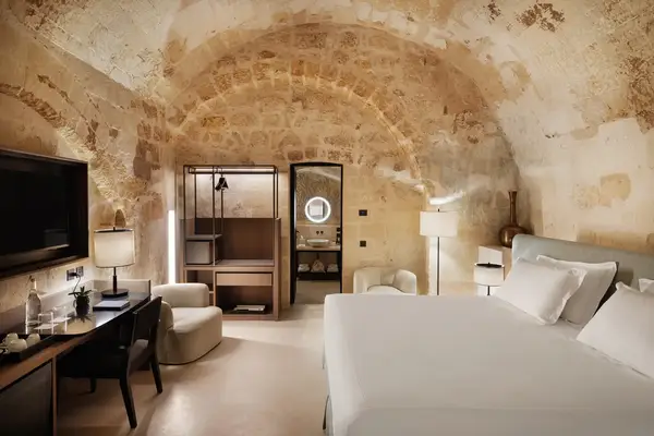 A bedroom with stone walls, a bed and nightstands, a sitting area, a large mirror, and a view into a bathroom