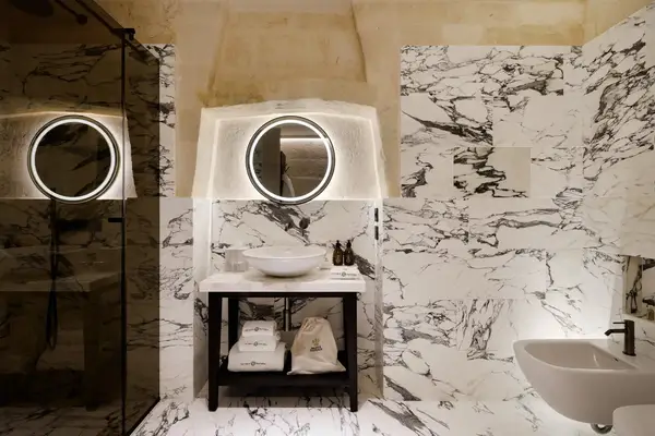 Marble bathroom featuring a lighted mirror above a vanity with a vessel sink, soap, and towels, a toilet and a glass shower enclosure