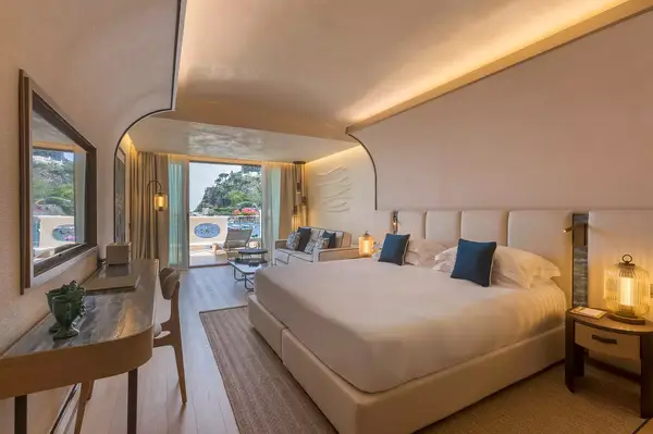 Hotel suite bedroom with a large bed, seating area, desk, and a balcony overlooking the sea in Italy