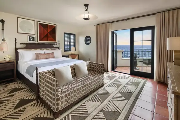 Hotel suite with a bed, seating area, and a window opening to a balcony with a view of the sea