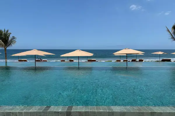 An infinity pool facing a beach with sun umbrellas and lounge chairs along the edge
