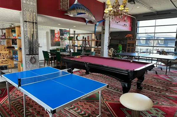 A game room featuring a table tennis table and a pool table, with decorative elements and furniture in the background
