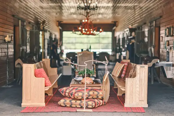 Rustic seating and decor in a stable setting with wooden benches and a central table with cushions and a chandelier above