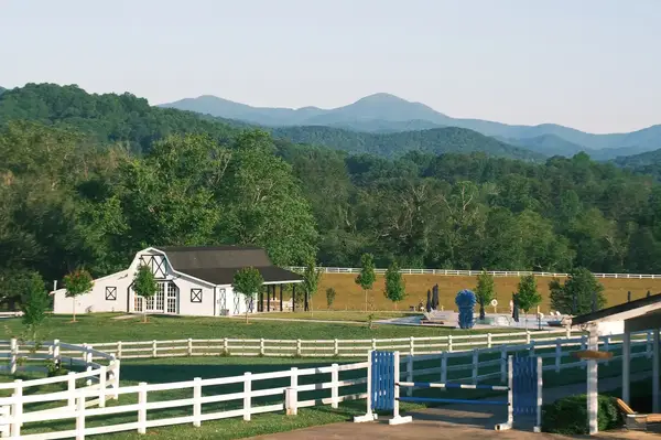 A scenic view of a farm with a barn, fenced fields, and a backdrop of green hills and mountains, horses visible in the area