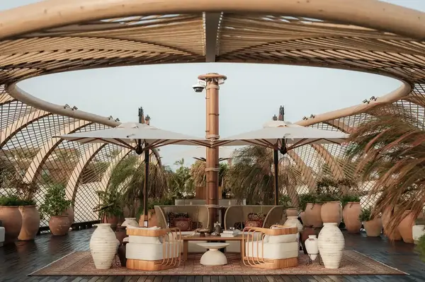 An outdoor lounge area with wicker furniture and umbrellas, under a wooden canopy surrounded by planters