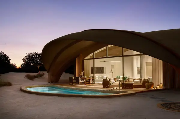 A luxury villa with a curved roof, a glass front, and an adjacent pool in a serene desert-like setting
