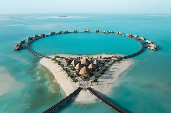 Aerial view of a coastal luxury resort with overwater villas forming a circular arrangement around a sandy island