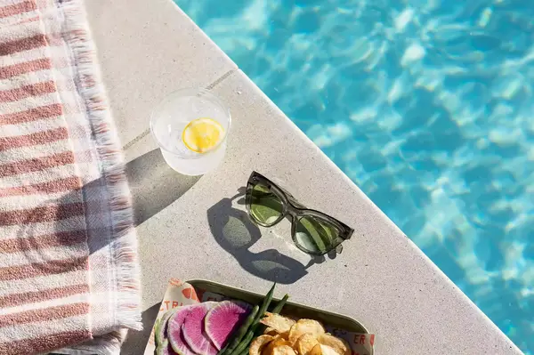 Poolside scene with sunglasses, a drink, and a plate of food on a ledge next to the water