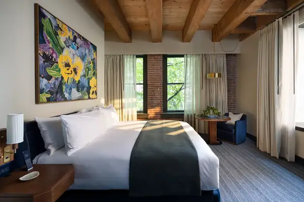 A hotel guest room with a bed, artwork, exposed wooden beams, and a window with curtains