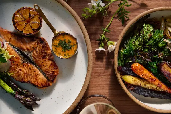 A plated fish dish with asparagus and a side of roasted vegetables on a wooden table
