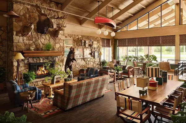 Spacious lodge interior with wooden beams, stone fireplace, hunting trophies, large windows, and casual seating and dining areas