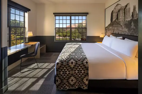 A modern hotel room with a king-size bed, geometric-patterned bedding, a desk, and a view of Sedona red rock formations through the windows