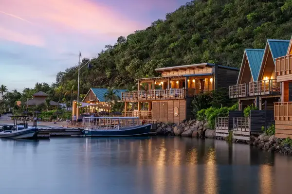 View of waterfront buildings with lit terraces and boats docked, situated against a lush green hillside at dusk