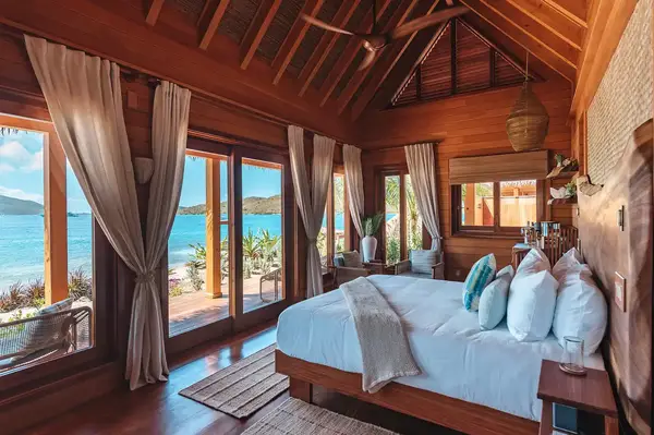 Interior of a beachfront bungalow with a view of the ocean through open doors, showing a bed, wooden furnishings, and curtains