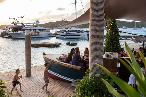 People dining at a waterfront venue with boats docked nearby, children playing on a wooden deck in the foreground