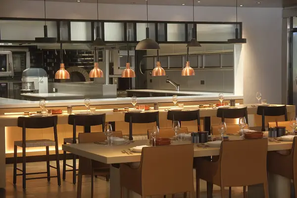 A dining area featuring tables, chairs, and overhead lighting, with an open kitchen in the background