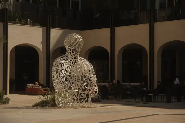 Outdoor courtyard with a large abstract statue in a seated pose, surrounded by an arched building