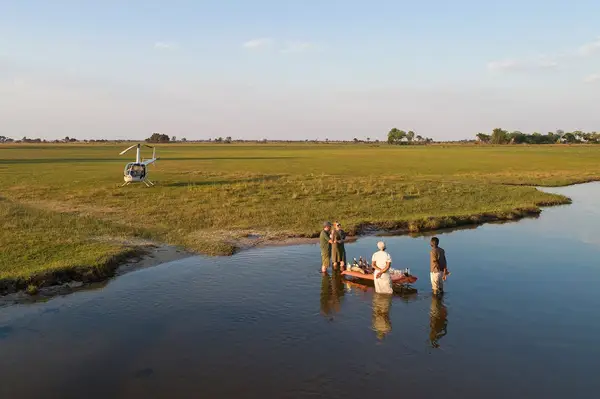 A helicopter sits in a grassy field while three individuals interact near a small boat in a shallow body of water