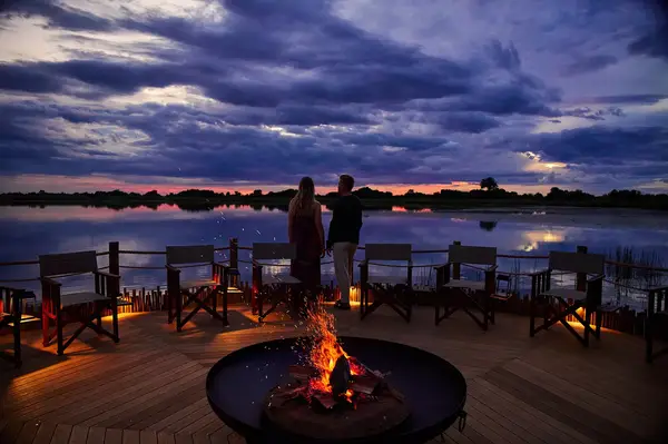 Two people stand on a deck with a fire pit, overlooking a serene body of water under a dramatic sky at sunset
