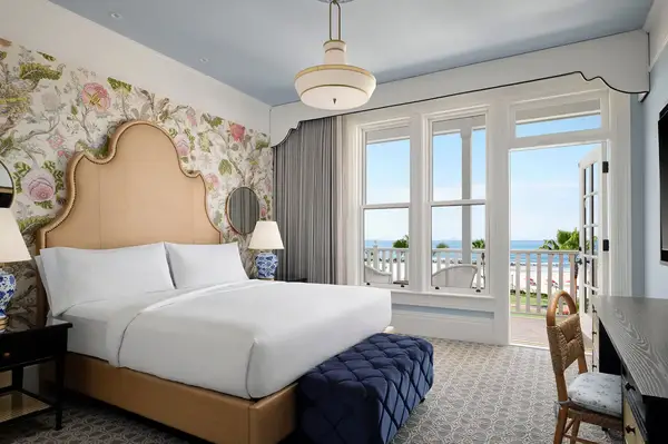 A stylish hotel room with a bed, floral-themed decor, a view of the balcony, and the ocean visible in the background