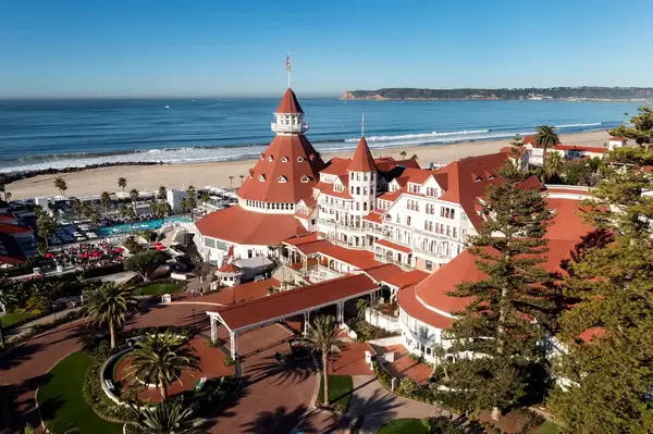 An aerial view of the historic Hotel del Coronado, situated by the Pacific Ocean, featuring its iconic red rooftops and surrounding palm trees