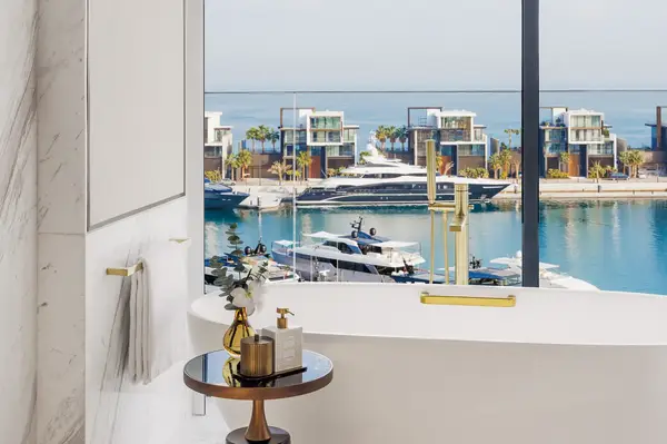 A luxurious bathroom with a bathtub by a large window overlooking a marina with yachts and modern waterfront villas