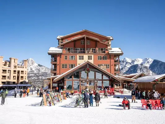 A ski lodge backed by mountains, beneath a blue sky, with people in the foreground