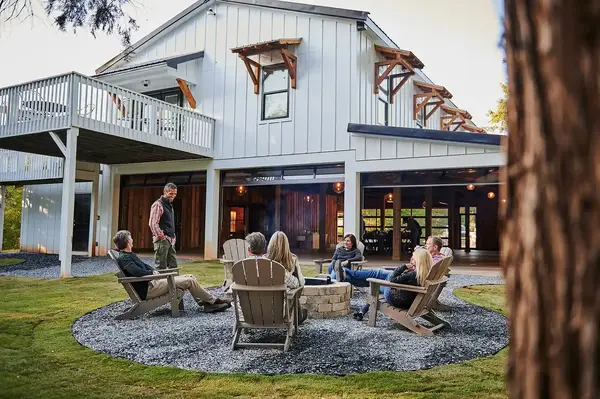 A group of people seated around a fire pit outside a rustic building with large windows and wooden accents