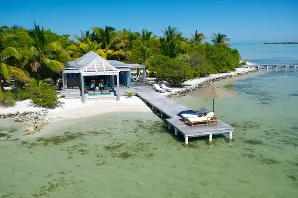 Tropical overwater bungalow with deck and lounge chairs, surrounded by clear shallow ocean waters and lush trees