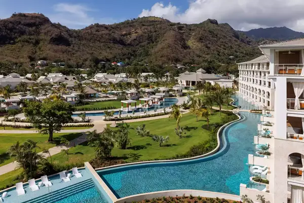 A resort with a meandering pool set against a mountainous backdrop and landscaped greenery, featuring multiple buildings and leisure areas
