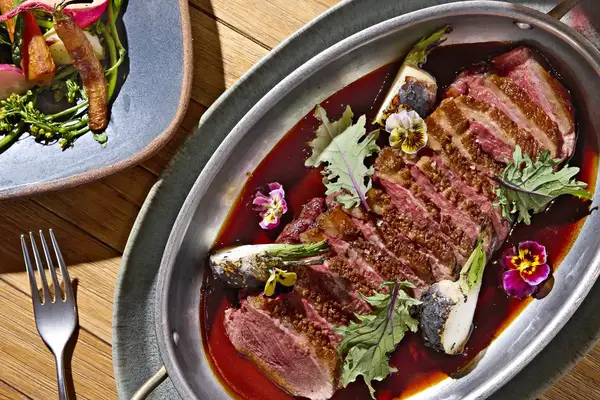 Gourmet plated dish with seared meat slices garnished with small flowers and greens alongside a fork and a side salad plate