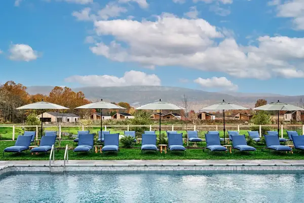 An outdoor pool with lounge chairs and umbrellas a grassy area and cottages in the background against a mountain backdrop