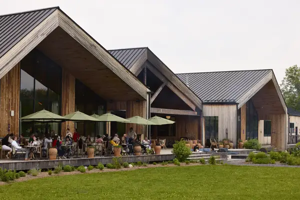 A wooden building with glass windows a patio with people seated under umbrellas surrounded by greenery