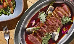 Gourmet plated dish with seared meat slices garnished with small flowers and greens alongside a fork and a side salad plate