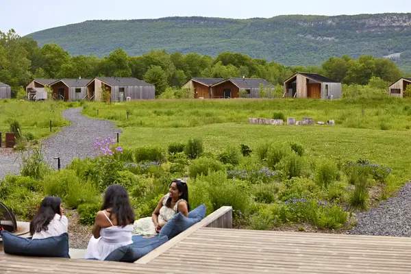 Three people sitting on a wooden deck overlooking cabins and a grassy landscape with hills in the background highlighting a peaceful rural setting
