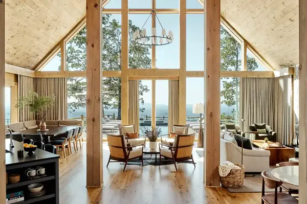 Interior of a luxury cabin with tall windows a lounge area and dining setup facing an outdoor deck with a scenic view