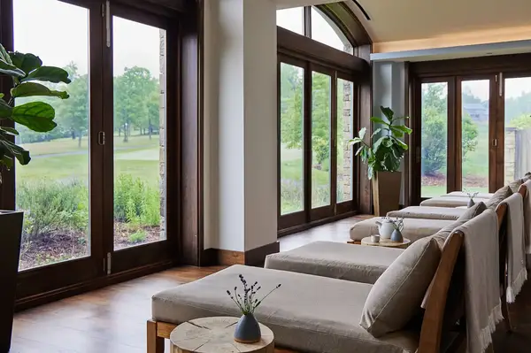 Relaxation room with lounge chairs plants and large woodenframed windows overlooking greenery