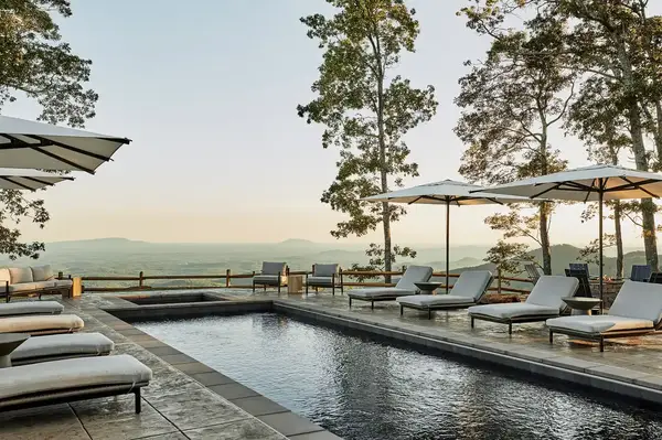 Outdoor pool area with lounge chairs and umbrellas surrounded by trees and overlooking a distant landscape