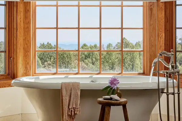 A freestanding bathtub in front of a window with a nature view folded towel and decor on a stool to the side faucet nearby