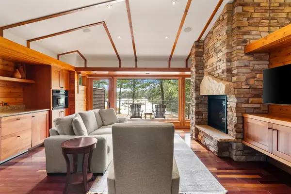 A cozy living room with wooden paneling a stone fireplace and large windows overlooking an outdoor patio and trees
