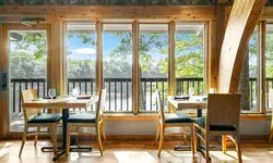 Restaurant seating area with wooden furniture and large windows overlooking a scenic outdoor view with trees and a body of water