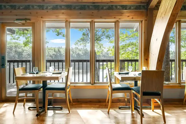 Restaurant seating area with wooden furniture and large windows overlooking a scenic outdoor view with trees and a body of water